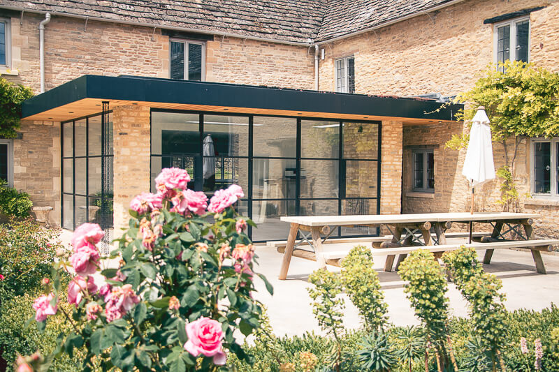 The rear open plan breakfast/dining room opens the house up to its grounds and brings in the light to enhance the interior. These traditional flush, double door, double glazed casement doors were custom made. They are made with slimline, leaded double glazing placed between the Cotswold stone pillars. The pillars themselves were taken out and re-built and they were re-formed to frame the new windows.
<br>
<br>
The roof flashing here was re-done in black to match the metal casement windows. The Japanese downpipe drain adds an interesting feature to the glazed corner, also enhanced by the downlighting  installed. A beautiful add on where new meets old but adding a hint of 'modern' touches . 
New parts of the patio were created to accommodate new drainage. The patio was re-laid in stone to complement the Cotswold stone building.
